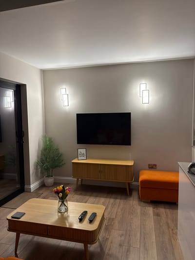 This image captures a modern and tidy living space, likely within a residence in Harrogate, United Kingdom, during the evening or night, as indicated by the active artificial lighting. The room features light grey walls and a warm, light wood-effect laminate floor.

In the center of the main wall, a large black flat-screen television is mounted. Below it, a stylish wooden TV cabinet with a distinctive slatted design provides a surface for decor. On this cabinet, a small framed sign is visible, bearing the text: "Is kettle broke?".

To the left of the TV cabinet, an artificial green plant in a white pot adds a natural element. Two modern, geometric light fixtures, composed of illuminated rectangles, are mounted on the wall above and below the television, providing bright, accent lighting.

On the right side of the image, an orange sofa or bench is positioned against the wall, near an electrical outlet. In the foreground, a rectangular wooden coffee table, matching the slatted design of the TV cabinet, holds a glass vase filled with colorful artificial tulips in pink, yellow, and purple. Two black remote controls and a dark coaster are also placed on the table.

A dark-framed door with glass panels is partially visible on the far left, reflecting the internal lights. The overall scene suggests a contemporary, well-maintained, and comfortable indoor setting. No people are present in the room.