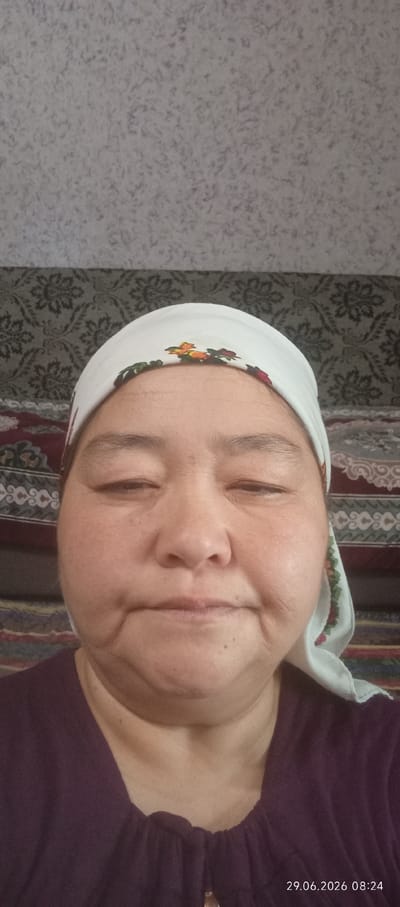 A woman is captured in a close-up, head-and-shoulders selfie. She is wearing a white headscarf adorned with colorful floral embroidery. Her eyes are closed, and her lips are slightly parted, giving the impression of a gentle smile or a relaxed state. The background appears to be indoors, with patterned wallpaper or fabric visible behind her. The lighting is soft, casting a natural glow on her face. The timestamp in the lower right corner indicates "29.06.2026 08:24," suggesting the image was taken on June 29, 2026, at 8:24 AM. The image appears to have been taken in the city of Chon-Kara, Kyrgyzstan.