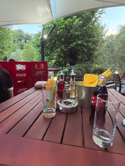 The image depicts an outdoor dining scene, possibly at a restaurant or cafe in Milanovo, Bulgaria. A wooden table is laden with various items, including a bottle of Coca-Cola with an empty glass placed over it, a metal napkin holder containing yellow napkins, a set of oil and vinegar dispensers, an ashtray, and a glass bottle of orange juice with a straw and an empty glass. Stacked Coca-Cola crates are positioned nearby. Trees and greenery are visible in the background, suggesting a location in or near nature. A partial view of a person's clothing (black shirt with white sleeve) is present.