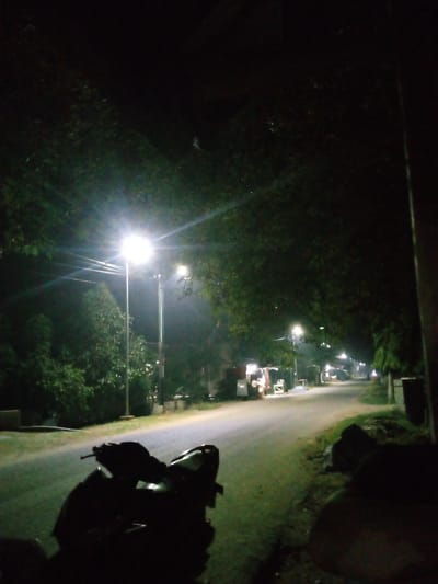 The image depicts a dimly lit street scene at night in Meunyelhee, Indonesia. A motorcycle is parked in the foreground, silhouetted against the pavement and surrounding vegetation. Streetlights illuminate the road, casting a yellowish glow on the asphalt and the trees that line the street. In the middle ground, there are several small roadside stalls, some appearing to be food vendors. The overall atmosphere is quiet and serene, with the artificial lights piercing the darkness.