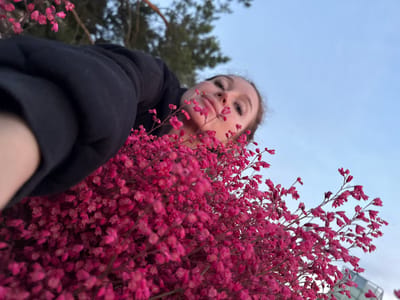 A low-angle, medium shot captures a young woman from the chest up, partially obscured by a cluster of vibrant pink flowers in the foreground. She is wearing a black hooded jacket. The woman is looking upwards, her face tilted towards the camera with her lips slightly parted. Her eyes are open, and her eyelashes are prominent. In the background, a clear blue sky is visible, with the suggestion of trees and possibly a building on the right edge of the frame. The image appears to have been taken outdoors, with natural light. The focus is on the woman and the flowers, with the background softly blurred. There is no indication of the specific location in Maksimovo, Russia.