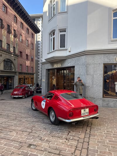 The image depicts a vibrant street scene in Sankt Moritz, Switzerland, on a clear day, centered around classic cars. In the foreground, a red classic Ferrari 275 GTB/4 is prominently positioned, viewed from its rear three-quarter angle on a cobblestone street. The vehicle bears an Italian license plate "MI 6U0765" and a white circular race number "34" on its left flank. Its chrome bumper and dual exhaust pipes are visible.

To the right of the Ferrari, a male individual, estimated to be between 40 and 60 years old, stands on the sidewalk. He wears a light olive-green jacket and sunglasses, holding a dark smartphone horizontally to photograph the Ferrari. He is located directly in front of the entrance to a Prada boutique, which occupies the ground floor of a light grey building. The Prada store features large display windows showcasing mannequins and merchandise. The building's lower facade is clad in grey, veined polished stone, while the upper sections are smooth light grey stucco with multiple windows.

Further down the cobblestone street, a red vintage car, possibly a Fiat Topolino Giardiniera, is visible, displaying a white circular race number "32" and a Swiss license plate "GR 10A 7H". Behind it, a dark-colored classic car with a visible white circular number is partially obscured. Several indistinct pedestrians are present in the background.

On the left side of the street, a multi-story building features a distinctive reddish-brown facade adorned with geometric patterns and multiple vertical friezes depicting figures. This building has numerous windows with dark frames, some featuring decorative ironwork. A sign for "CONDITOREI HANSELMANN" is visible on a ground-floor shop. The street itself is paved entirely with light-colored, rectangular cobblestones. The scene is illuminated by bright sunlight, casting distinct shadows on the building facades and the street surface. A clear blue sky is visible above the buildings.