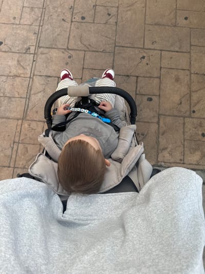 An overhead shot captures a child sitting in a stroller, facing away from the camera. The child is wearing a gray shirt, striped pants, and red sneakers. A pacifier clip with letter beads is attached to their clothing. The stroller features a black handle and a beige-colored interior. The stroller is being pushed, as a gray hooded sweatshirt can be seen close to the stroller, presumably worn by the person pushing it. The stroller is on a paved walkway made of square tiles.