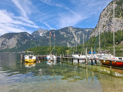 In Obertraun, Austria, a tranquil scene unfolds at a lakeside dock. The crystal-clear water of Hallstätter See reveals a rocky bottom and reflects the serene atmosphere. Several sailboats, with their masts reaching towards a sky dotted with wispy clouds, are moored to a wooden pier. Among them, a bright yellow motorboat is prominently docked, its stern facing the viewer. The backdrop is a majestic mountain range covered in dense green forest, with rocky outcrops interspersed amongst the trees. The image captures a peaceful moment of leisure and natural beauty in this Austrian town.