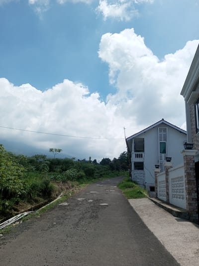 The image depicts a slightly potholed asphalt road leading uphill between vegetation on the left and two white houses on the right, under a cloudy sky. On the left, lush green plants and trees line a shallow ditch. On the right, the first house is a two-story structure with a balcony and decorative windows. Adjacent to it is a fence with alternating brick and concrete sections, and the second house. A power line runs across the top of the frame. In the distance, mountains are visible through the vegetation. Kioumaknao 2, Laos is not identifiable from this image.