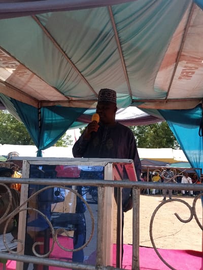 A man in a grey robe and a red and black hat is speaking into a microphone at a podium. The podium is located under a canopy tent with a blue top and a white bottom. The man is standing in front of a crowd of people gathered in a dusty area. The canopy is supported by metal poles, and there are trees in the background. The location is Zum, Nigeria, and the coordinates are 10.116, 13.046. The man is looking at the audience. He is holding the microphone in his right hand and his left hand is resting on the podium. A metal railing with a curly design is positioned in the foreground. The railing is painted black. The audience is sitting on chairs and standing in a casual manner. There is a red carpet on the ground in front of the podium. The man is speaking into the microphone and seems to be engaging with the audience.