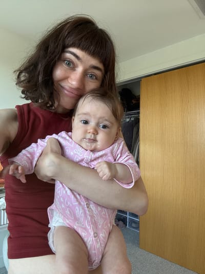 A woman with brown hair and green eyes is holding a baby in a pink onesie. The baby has food around its mouth. They are inside, likely in a bedroom, as a closet with clothes and a shoe rack is visible in the background. The location is Springfield, United States of America.