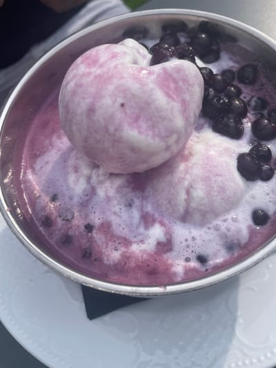 A close-up, overhead shot shows a metallic bowl filled with a vibrant purple liquid, topped with two scoops of light pink and white swirled ice cream. Whole blueberries are scattered throughout the liquid and around the ice cream, some appearing slightly submerged. The liquid is frothy with small bubbles, particularly around the edges of the bowl and the ice cream. The bowl itself is round and metallic with a visible rim. The bowl rests on a white, textured plate, which is then placed on a surface that is not fully visible but appears to be light-colored. A black napkin is tucked under the rim of the plate on the right side. The background is blurred, but suggests an outdoor setting with hints of natural light and possibly the presence of other people, one of whom is partially visible in the top left corner. The setting is likely Alagna Valsesia, Italy, given the context.