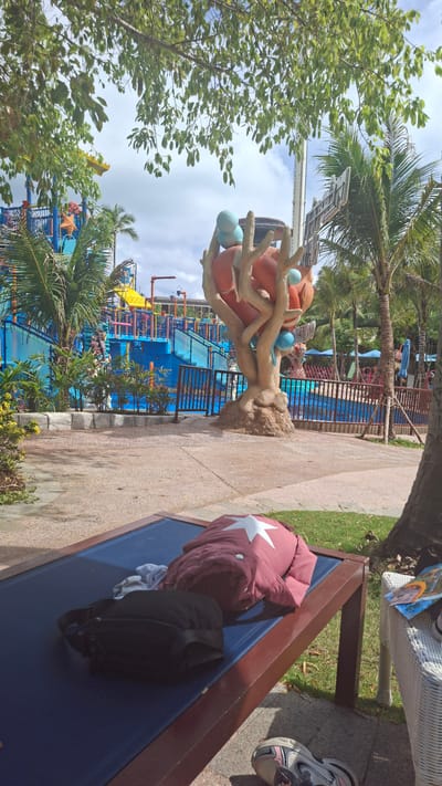The image depicts an outdoor scene in Phú Quốc, Vietnam, possibly at a water park or resort. A colorful water park structure with slides, pools, and themed elements like starfish and palm trees is visible in the background. A faux tree sculpture with orange spheres and blue orbs stands in the foreground. The tree is situated near a paved walkway. A blue bench with a burgundy jacket featuring a white star and a black bag resting on it is partially visible on the right side of the frame. Trees are present on the left side, partially obscuring the view.