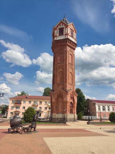 The image captures a detailed view of the Staraya Russa Water Tower, a prominent red brick structure, under a bright blue sky with scattered white cumulus clouds. The tower stands centrally in the frame, exhibiting a tall, slender form with decorative arched elements along its shaft. Its upper section features a distinct octagonal observation level with windows, detailed white and orange-red trim, and a pointed, multi-faceted dark gray roof culminating in a small spire.

In the foreground, slightly to the left of the tower, a dark metal statue depicts a man pushing a two-wheeled cart carrying a large barrel. A dog walks faithfully beside the man, who is rendered in period clothing including a hat and a long coat. The ground in the immediate vicinity is paved with geometric patterns of red, brown, and light beige interlocking tiles.

To the left of the water tower, a multi-story building with a light peach-colored facade and a red tiled roof is visible. It features several windows and balconies with dark railings. An inscription "ЖЕМЧУЖИНА" (ZHEMCHUZHINA) is visible on its lower level. Further left, a street with an "A" pedestrian crossing sign and faint figures of pedestrians can be discerned. To the right of the water tower, a portion of another building with a white facade, red roof, and arched windows is partially visible, along with dark, ornate streetlights. Green trees and shrubs provide additional landscaping around the buildings.
