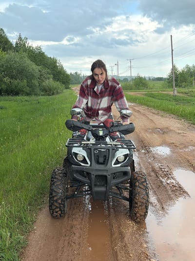 A female individual is on a black and white ATV. She is on a muddy dirt path. The landscape includes green grass, trees, and cloudy skies. Power lines are visible in the background. The location is in Nizhnii Vishur, Russia.