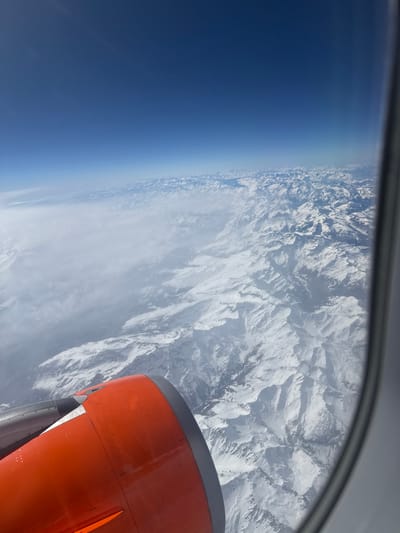 The image presents an aerial view captured from an aircraft window, looking down upon an extensive snow-covered mountainous landscape. In the lower-left quadrant, a significant portion of an aircraft engine nacelle is visible; it is predominantly bright orange-red with a dark grey or silver leading edge. A metallic grey component, likely part of the engine pylon or wing, is situated above the orange nacelle. The right side of the frame reveals a segment of the aircraft window frame.

Below the aircraft, vast ranges of rugged, snow-covered mountains stretch into the distance. These mountains feature prominent peaks, deep valleys, and shadowed slopes, indicating significant topographical variation. A diffuse layer of white clouds partially obscures the mountains in the mid-left and central portions of the scene, creating areas of reduced visibility. Above the terrain and clouds, the upper two-fifths of the image consists of a clear, deep blue sky, fading to a lighter hue near the horizon line. The scene is illuminated by bright daylight, casting distinct shadows across the snow-covered peaks.

This mountainous terrain is consistent with the Swiss Alps, specifically the region surrounding the city of Bad Ragaz, Switzerland. From the depicted high altitude, specific urban features of Bad Ragaz are not discernible; the view encompasses the broader, snow-covered Alpine environment. No people or specific interactions are visible within the frame.