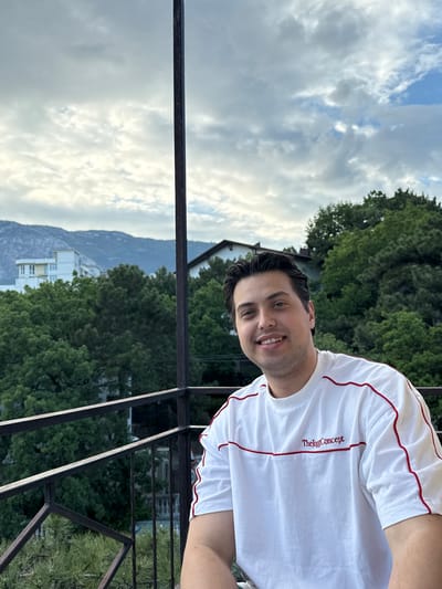 A man stands on a balcony in the city of Кореизский поселковый совет, Ukraine. He is smiling and wearing a white t-shirt with red trim and the word "Thekggconcept" in red lettering. Behind him is a landscape view of trees, a mountain, buildings, and a cloudy sky. The balcony has a dark metal railing.