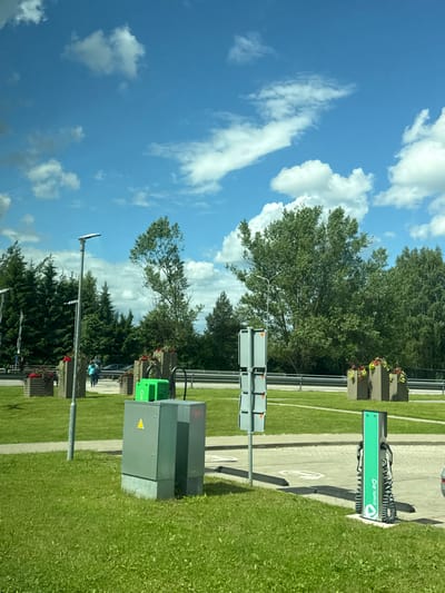 The image depicts an outdoor scene in Jaunolaine, Latvia, under a bright blue sky with scattered white clouds, suggesting daytime and fair weather. The perspective is from a low angle, looking upwards.
The main subjects include a street light, electrical equipment, and an electric vehicle charging station. The charging station is a prominent green unit with the text "Enefit" and a logo visible. Next to it is a gray electrical cabinet with a yellow triangular warning symbol. In the background, there are trees, some with planter boxes containing flowers. There are also paved pathways and a grassy area.
A few people are visible in the background, appearing to be walking or standing near the charging area. One person is wearing a blue shirt, and another is wearing a light-colored top. They seem to be engaged in casual activity, perhaps waiting or interacting.
The scene appears to be a public space, possibly a parking lot or a service area, equipped for electric vehicle charging. The overall atmosphere is calm and ordinary, with no specific events or strong emotions conveyed. The presence of the charging station and electrical equipment suggests a modern, functional setting.