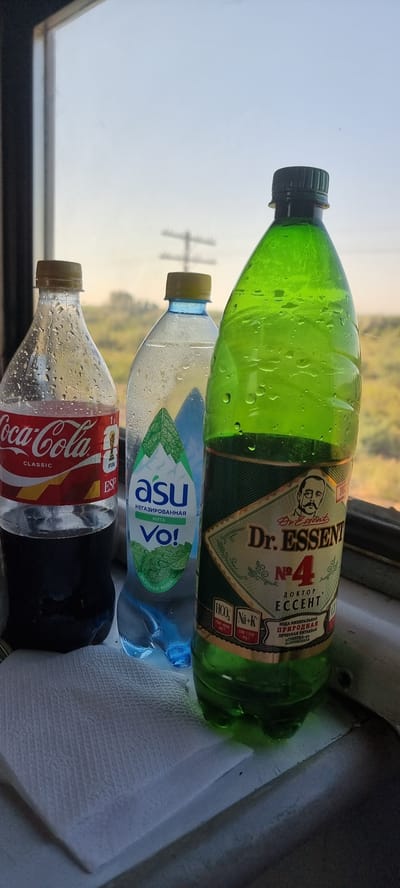 This media file shows a close-up, rotated view of three plastic beverage bottles placed on a white cloth or napkin. The bottles are stacked vertically. The topmost bottle is a vibrant green and appears to contain a carbonated drink, possibly with a label indicating "Dr. Essent" and "№4". The middle bottle is clear and contains a light blue liquid, with a label that reads "asu Vo!" and mentions "негазированная мята" (non-carbonated mint) in Russian. The bottom bottle is a clear Coca-Cola Classic bottle, identifiable by its iconic red label with white lettering. All bottles are covered in condensation, suggesting they are cold.

In the background, the scene appears to be a train window, indicated by the presence of a window frame and a blurry view of the outdoors. The outdoor scene is bright and shows trees and power lines, suggesting it is daytime. The lighting is soft, hinting at either early morning or late afternoon. The overall mood is casual, possibly capturing a moment during travel. The text on the bottles includes brand names and descriptive information about the contents.