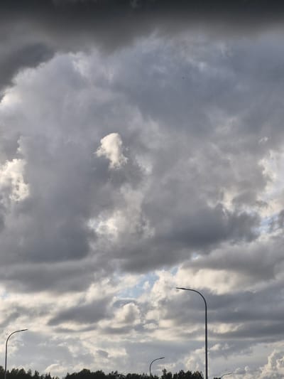 The image depicts a cloudy sky over a landscape that includes a treeline and streetlights, indicative of an urban or suburban environment. The clouds are a mix of dark gray and lighter white, suggesting a dynamic weather system. The treeline at the bottom suggests vegetation, possibly a park or a natural border within the city. Several streetlights with curved arms are visible, implying a road or public area. The overall atmosphere is somber due to the overcast conditions. There are no people or specific actions or interactions captured in the frame. The scene can be considered a generic view of an outdoor environment in Jenči, Latvia, under cloudy skies.