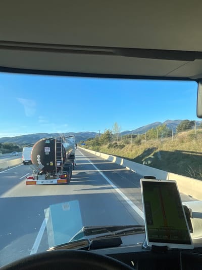 The image depicts a forward-facing view from the interior of a vehicle, likely a truck, traveling on a multi-lane highway near Maureillas-las-Illas, France. A clear blue sky is visible above. In the foreground, a portion of the vehicle's dashboard and steering wheel is present, with a white tablet device mounted vertically on the right side of the dashboard, displaying a green-hued navigation map. Directly ahead, a black tanker truck with a silver cylindrical tank is positioned in the right lane, featuring an "80" speed limit sticker and hazard placards on its rear. Reflections of the tanker truck and the sky are visible on the vehicle's windshield. A white van and another light-colored vehicle are visible in the left lane further down the highway. The roadway is marked with white solid and dashed lines. To the right of the highway, a light-colored concrete barrier separates the road from a grassy embankment with scattered trees and a wire fence. Mountains or hills are visible on the horizon. Long shadows are cast across the highway from the barrier and vehicles, indicating a low sun angle.