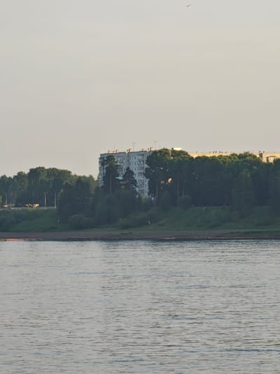 The image shows a wide river in the foreground, with calm, rippling water reflecting the pale sky. The riverbank on the far side is lined with a dense growth of dark green trees, interspersed with taller pine trees. Behind the trees, a multi-story, light-colored apartment building with a long, horizontal structure is visible. The building has numerous windows and appears to be in the city of Lesosibirsk, Russia. The sky is overcast and a light, hazy color, with a faint, distant object, possibly an airplane, visible in the upper portion of the frame. The overall impression is of a natural landscape bordering an urban residential area.
