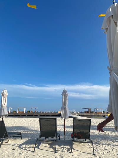 The image shows a beach scene in Marina Maroma, Mexico. Three closed, white beach umbrellas are present along the sand, with accompanying dark-colored beach chairs arranged in rows. The sky is predominantly clear blue with streaks of white clouds. A hand is partially visible at the upper right edge of the image. A yellow object is dangling in the sky. The beach appears to be lined with structures and vegetation in the background.