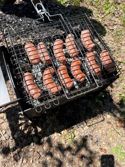 A close-up, high-angle shot shows ten plump, reddish-brown sausages cooking on a black wire grill basket over embers in a portable charcoal grill. The grill is situated on a ground covered with small pebbles and dirt, with some sparse green vegetation visible in the background. The sausages appear to be in various stages of cooking, with some showing slight browning and charring. The lighting suggests it is a sunny day, casting shadows from the grill and some nearby foliage onto the ground. This scene could be anywhere with outdoor grilling, including the city of Ishimbay, Russia, where it might be a common recreational activity in parks or backyards.