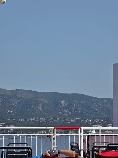 This is a vertical, medium-shot photograph taken from a ferry in Magaluf, Spain, looking inland towards a mountain range. The sky is clear and blue, taking up approximately two-thirds of the frame. In the foreground, a white metal railing with a red top edge separates the ferry deck from the water below. To the left, a black metal table with two black chairs is visible. One chair has "ESTRELLA DAMM" printed on its back. A person's legs and torso are visible lying down on the right side of the frame, wearing a blue shirt and a navy blue baseball cap with a white "NY" logo. Behind the railing and in the middle ground, a body of water is visible. Beyond the water, a densely vegetated mountain range dominates the landscape, with a town or village nestled at its base, characterized by numerous buildings. The overall scene suggests a relaxed atmosphere, possibly on a sunny day during a ferry journey.