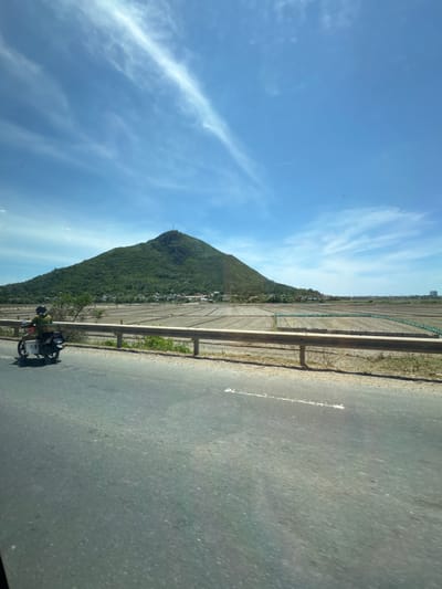 The image depicts a landscape scene near Tuy Hoa, Vietnam, featuring a prominent, tree-covered mountain in the background. In the foreground, a paved road runs horizontally, with a motorcycle carrying a single rider traveling from left to right. The motorcycle has a white box attached to its rear rack. Adjacent to the road is a field that appears to be a cultivated area, possibly for agriculture, with a visible guardrail separating it from the road. The sky above is a clear blue with wispy white clouds, indicating a daytime setting. A few buildings are visible in the distance, clustered around the base of the mountain and extending towards the right side of the image. The overall impression is one of a rural or semi-rural environment under a bright sky.