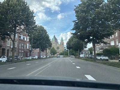 The image shows a street scene in Maastricht, Netherlands, with a large church visible in the distance. The street is paved with asphalt and has white lane markings. There are trees lining the street on both sides, with some residential buildings also visible. Several cars are parked along the side of the road and driving on the street. The sky is partly cloudy. The church in the background has a large circular window and two spires.