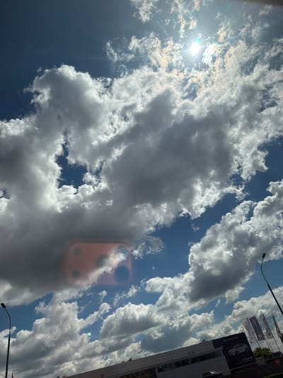 The image shows a bright, partly cloudy sky over Dzerzhinsky, Russia. The sun is visible, shining through gaps in the clouds, creating lens flares on the camera. Below the sky, a building with a banner displaying "CHANGAN AUTO" and images of cars is visible. Several flags with the "CHANGAN" logo are also present. A street lamp is positioned to the right of the frame, and a portion of what appears to be a vehicle is visible at the bottom of the image. A pink, rectangular object with visible camera lenses is reflected or superimposed onto the clouds in the middle of the frame. The environment suggests an outdoor commercial area, likely a car dealership.