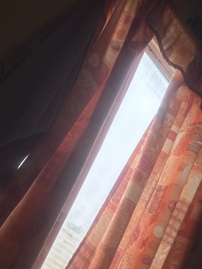 The image displays an interior view of a room in Alderbury, United Kingdom, looking outward through a window. Two drawn curtains, exhibiting a patterned design in hues of orange, red, and beige with circular, square, and leaf motifs, frame a central vertical window opening. The curtains are pleated, with the left panel obscuring a dark, textured interior surface on the far left of the frame. Through the window, a bright, overcast or hazy sky is visible, with a faint, indistinct outline of a distant landscape or structures through the translucent haze. The window pane exhibits minor smudges and dark particulates. No individuals or specific actions are depicted.