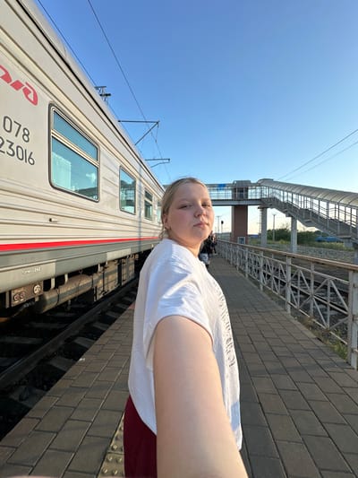 A selfie-style photograph, taken on a bright, clear day, captures a young person standing on a train station platform. The image is oriented vertically with the sky on the left and the train tracks on the right, as if the camera was held sideways.
The main subject is a person with light-colored hair, wearing a white t-shirt and what appears to be dark red or maroon shorts, facing the camera with a neutral expression. Their left arm and shoulder are visible, extending towards the camera.
To their right, a beige or light gray train car is prominently featured, parked at the platform. The train displays the red "РЖД" (RZD - Russian Railways) logo, along with the numbers "078" and "23016" on its side. Below the train windows, a distinct red stripe runs horizontally. Parts of the train's undercarriage, including what looks like an air tank and other mechanical components with the text "ТОРМОЗ 242" (BRAKE 242), are visible between the train car and the platform.
The platform itself is paved with dark gray rectangular pavers. Beyond the train, railway tracks are visible, and in the background, an overhead metal pedestrian bridge or station structure spans across the tracks. Overhead electrical wires (catenary system) are also visible against the clear, blue sky. The sunny conditions are evident from the bright illumination and distinct shadows. The overall setting is an active railway environment, likely in Zimovniki, Russia, given the "РЖД" branding.