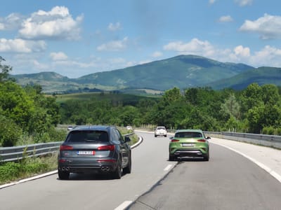 This media file depicts a scenic drive on a sunny day in Osikovitsa, Bulgaria. The scene is a paved road curving through rolling green hills and lush forests. In the foreground, two SUVs are driving away from the camera. The one on the left is a dark grey Audi, and the one on the right is a bright green Kia. Both vehicles are captured from the rear, showcasing their taillights and license plates. The Audi's license plate reads "830 M 358". The Kia's license plate is "EH 4901 MA".
Further down the road, other vehicles are visible, including a white SUV and a dark car in the distance. The sky is a vibrant blue, dotted with fluffy white clouds, suggesting pleasant weather. The surrounding landscape is characterized by verdant vegetation, with trees lining the sides of the road and hills stretching into the background. The time of day appears to be midday, given the bright sunlight casting shadows. The overall mood is one of leisurely travel and appreciation for the natural beauty of the Bulgarian countryside.