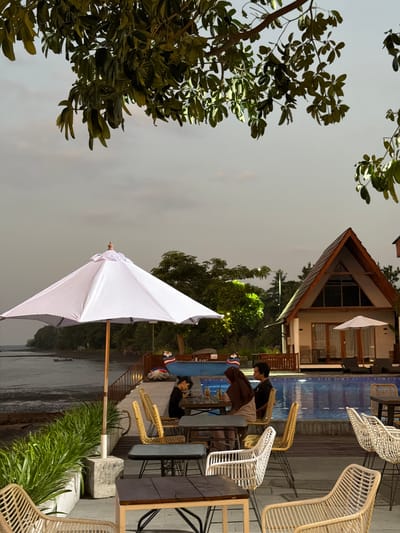 The image depicts an outdoor seating area with a swimming pool and a body of water in the background, likely taken at dusk or night given the lighting. Two individuals are visible. One person, wearing a black shirt and sunglasses, is seated at a table and looking at their phone. Another person, wearing a brown jacket over a white shirt, is also seated at the same table, engaged in conversation with the first individual. Both are seated on rattan chairs.
In the foreground, a large white umbrella is partially visible, suggesting it might be an outdoor cafe or restaurant setting. The surrounding vegetation, including trees with dark green leaves, adds to the tropical ambiance. In the background, a modern building with a triangular roof and large windows is illuminated, indicating it might be a resort or hotel. Rattan furniture, including chairs and lounge seats, is arranged around the pool area. The sky appears overcast, and the overall mood is relaxed and serene. The location context points to Pasir Putih, Indonesia.