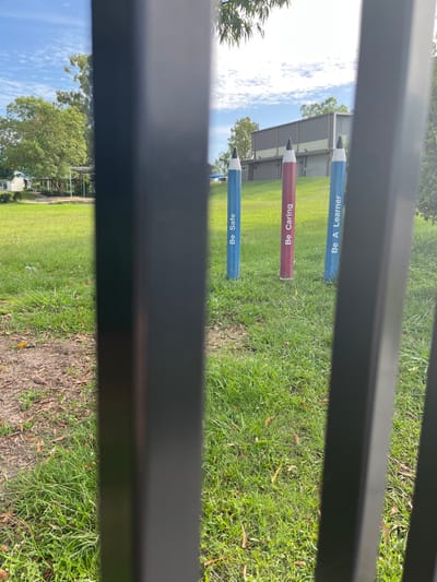 The image displays an outdoor scene in Riverhills, Australia, viewed through the vertical bars of a dark metal fence. Three large, colorful, pencil-shaped markers are prominently positioned upright in a sunny, green grassy field. The sun is high in the sky, suggesting midday or early afternoon, with clear skies and scattered white clouds.

From left to right, the first pencil is blue and bears the text "Be Safe". The middle pencil is red and inscribed with "Be Caring". The third pencil, also blue, reads "Be A Learner". These markers are planted in a well-maintained lawn, which shows some patches of dry grass and scattered leaves.

In the background, a low-rise building with a grey roof and light-colored walls is visible, possibly a school or community facility. Lush green trees flank the building against the bright blue sky. The scene conveys a sense of a community or educational environment, emphasizing positive behavioral values. No individuals are present in the image.