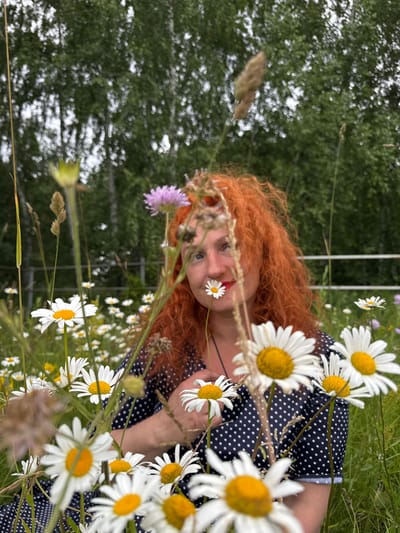 A woman with bright red, curly hair is partially visible behind a field of white daisies in Privolzhsky, Russia. She is wearing a dark blue dress with white polka dots and has a single daisy positioned in front of her mouth. The background is softly blurred, showing trees and a white fence. The focus of the image is on the woman and the surrounding flowers, suggesting a natural, outdoor setting.
