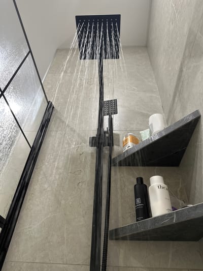 In a modern shower stall with light gray marble-patterned tile walls, a square black showerhead is actively spraying water downwards. A black shower fixture extends vertically along the wall. To the left, a partial view of a glass shower door with black framing is visible. Two dark gray, triangular shelves are integrated into the wall. The upper shelf holds a white bottle, a small green and white patterned sachet, and an orange-labeled bottle. The lower shelf displays a black bottle labeled "DS Skabaro" and a white bottle with the text "the act EXTRA". The scene depicts the functionality of a shower in a private bathroom setting.