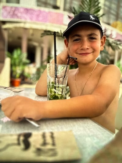 A young boy, likely under 10 years old, is captured in a candid, slightly tilted, close-up shot. He is wearing a black baseball cap with a white logo on the front and a thin gold chain around his neck. The boy has rosy cheeks and a gentle smile, looking towards the camera with his head resting on his hand. He is holding a straw to his lips, which is inserted into a tall glass filled with a green beverage and ice. The background is blurred, suggesting an outdoor setting. Hints of green foliage and what appear to be pink and white striped fabric or structures are visible, creating a summery, relaxed atmosphere. The lighting suggests it is daytime, possibly late afternoon. The boy's expression conveys happiness and contentment. No visible text is present in the image.