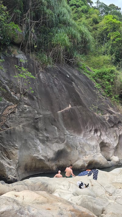 The image depicts a natural outdoor scene in Nam Ninh Hoa, Vietnam, dominated by large rock formations and lush vegetation. A cliff face with a mix of dark, smooth rock and areas covered in green foliage occupies a significant portion of the left side. In the upper left corner, dense trees indicate a forested area. To the right, light-colored rock formations create a pool of water. Two shirtless individuals are positioned near the edge of the pool, appearing to be either swimming or preparing to enter the water. Clothes and other personal belongings are placed on a rocky ledge adjacent to the pool.