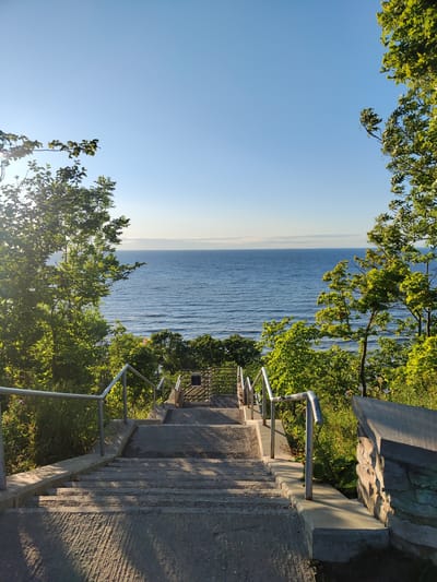 This image shows a concrete staircase descending towards a body of water, likely a lake or ocean, under a clear blue sky. The staircase is flanked by lush green trees and foliage on either side, with sunlight casting shadows on the steps. Metal handrails run along both sides of the staircase, with stone accents on the right. In the distance, the water is calm with subtle ripples, meeting a distant horizon with a faint line of clouds. The scene is devoid of any people or visible structures beyond the immediate staircase and its surroundings. The location is identified as Valaste, Estonia, suggesting this is likely a scenic overlook or access point to the coast.