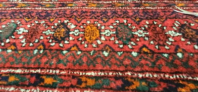 The image shows a close-up view of a patterned rug. The rug has a predominantly red base with intricate geometric designs in various colors, including dark blue, green, orange, and white. The pattern includes zig-zag lines, diamond shapes, and circular motifs. The texture of the rug appears to be woven or knotted, with visible threads and a slightly fuzzy surface. In the upper right corner of the image, a portion of a silver object with two pointed prongs is visible, suggesting it might be a utensil or a tool. The lighting is even, indicating it's likely taken indoors with artificial light or diffused daylight. There is no visible text in the image. The overall impression is that of a detailed textile with a traditional or ethnic design, possibly from a home or a market setting in Lod, Israel.