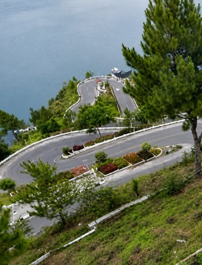The image shows a winding road with a switchback design, carved into a green, grassy hillside overlooking a large body of water. Pine trees are visible along the roadside and in the foreground. The road has white lane markings and concrete barriers along its edges. On the roadside, there are well-maintained flower beds with red, green, and orange foliage. In the distance, a white boat is docked near a dark-colored, umbrella-like structure. The overall scene depicts a scenic viewpoint or resort area, possibly part of the location identified as Turpuk Sihotang, Indonesia. No people are visible in the image.