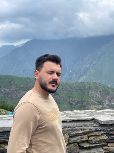 A man with a beard and dark hair stands in the foreground with his eyes closed, facing slightly to the right. He is wearing a light brown sweatshirt with a golden rectangular design on the left chest. Behind him, a stone wall or barrier is visible, constructed from irregular gray and brown stones. Beyond the wall, a lush green mountainside is illuminated by daylight, with a dramatic, cloudy sky overhead. The distant mountains are shrouded in a hazy, atmospheric perspective. The overall scene suggests an outdoor location, likely in a mountainous region, with hints of a remote or natural setting. The presence of a blue tarp and a fence on the left side of the frame suggest a possible man-made structure or a viewing area in the landscape.