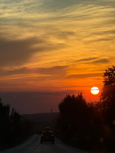 This is a landscape photograph taken at sunset in Zgalevo, Bulgaria. The image features a dramatic sky filled with vibrant orange and yellow clouds, streaks of lighter clouds are visible in the distance. The sun, a bright, glowing orb, is partially obscured by dark foliage in the foreground. A dark-colored SUV is driving on a road in the lower right portion of the frame. The scene evokes a sense of peace and the beauty of nature during the golden hour.
