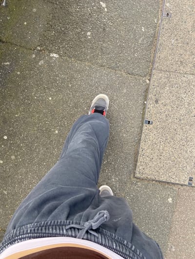 The image, taken in Broxbourne, United Kingdom, presents a high-angle view of an individual's lower torso and legs. The subject is wearing dark grey sweatpants featuring an elasticated waistband with a visible grey drawstring knot, and grey athletic shoes with white soles and orange or red accents on the visible right shoe's upper. A white band, possibly a garment hem, is visible beneath the sweatpants' waistband, partially revealing a brown or tan layer. The person is standing on an outdoor surface composed primarily of dark grey, granular asphalt or concrete pavement, exhibiting minor cracks and lighter specks. To the right side of the frame, a lighter grey, rectangular concrete utility cover with a metallic frame and two circular lifting points is embedded in the pavement.