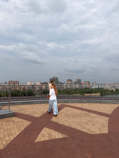 An overcast, cloudy sky looms over a city skyline and a waterfront. Two individuals are on a paved rooftop observation deck, which features a geometric pattern of tan and reddish-brown bricks. A metal railing separates the deck from a green treeline and a body of water below. In the distance, various multi-story buildings, including residential complexes and what appears to be a stadium or arena, are visible against the hazy horizon. One person, facing away from the camera, is wearing a white t-shirt and loose-fitting light blue pants, carrying a brown purse over their shoulder. Another person is partially visible, also facing away, wearing a light-colored top. The overall scene suggests an urban landscape with a focus on an elevated viewpoint.