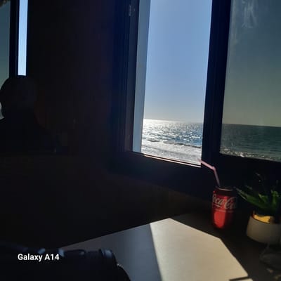 The image depicts a partial view of what appears to be a window or skylight offering a glimpse of a coastal scene. The foreground is dominated by a dark interior, possibly a room or building, with the edge of the window frame prominently visible. Outside the window, there is a view of the ocean with visible waves, and a distant shoreline with what could be buildings or structures, suggesting the location is near a populated coastal area. A red Coca-Cola can with a pink straw is positioned on a surface near the window, along with a potted plant. The image has the imprint "Galaxy A14" indicating it was taken by a Samsung Galaxy A14 mobile phone. The location is specified as Al-Mawasi, Unknown.