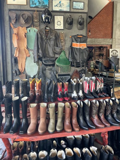 This image captures the interior of a retail establishment in Ban Yid, Thailand, displaying a curated collection of leather goods and Western/biker accessories. No individuals are present in the scene. The upper section features a black metal grid panel on which various items are mounted: a tan cowboy hat adorned with a metal badge, a large, flattened, tan crocodile or alligator skin, a large irregular piece of light green leather or hide, a black motorcycle helmet, a dark brown leather motorcycle jacket, and a black leather vest featuring an orange stripe with a "Harley-Davidson Motorcycles" logo. Assorted leather pouches, gloves, and chaps are also visible on the grid. Above the display, several framed pictures depicting horses with riders and motorcycles are affixed to a gray wall. To the right, an exposed red brick wall with a diagonal architectural element and a single suspended light bulb is visible.

The lower half of the image presents multiple wooden shelves arranged with rows of leather boots. The top shelf displays an assortment of highly decorative cowboy boots, including pairs with intricate yellow and red designs, black and white patterns, red and black motifs, and one pair featuring the American flag design with red and white stripes and white stars on blue. The middle shelf showcases multiple pairs of pull-on boots in various solid colors, predominantly black, different shades of brown, and olive green. The bottom shelf continues the display with numerous dark-colored cowboy and riding boots, many with visible decorative stitching or patterns, some containing white packing material within their shafts. The overall environment suggests a rustic or industrial-themed retail space specializing in custom or themed leather apparel.