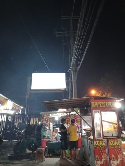 At night in Sekopek Wetan, Indonesia, a street food vendor operates a "Kong Fried Chicken" stall. Two individuals, possibly customers, are interacting with the vendor who is preparing food. The stall is illuminated by artificial lights, highlighting fried chicken being cooked and displayed. Behind the stall, a large blank white billboard is mounted on a yellow and blue structure. A street lamp shines brightly on the right side of the scene. Power lines are visible overhead, strung across the dark sky. The area appears to be a roadside market or commercial area, with motorcycles parked nearby and other structures suggesting ongoing activity.