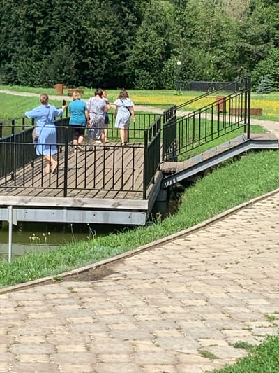 This image shows a group of four women walking on a wooden pier with a black metal railing. One woman in the foreground is wearing a light blue dress and holding a phone. Behind her, three other women are walking together, one in a teal shirt, another in a patterned dress, and the last in a light blue patterned dress. The pier extends over a body of water with grass and reeds on the bank. In the background, there are trees, a paved walkway, and a grassy area with some yellow flowers. The scene appears to be a park or recreational area on a sunny day. The location is not specified within the image or the prompt.