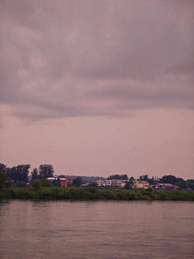 The image depicts a tranquil river scene with a distant village on the opposite bank. The sky is overcast with muted, purplish clouds. The river in the foreground is calm, with subtle ripples reflecting the soft light. A dense line of green foliage, likely trees and bushes, separates the river from the village. The village itself is comprised of several buildings, including a prominent red structure and lighter-colored buildings with red roofs. The architecture suggests a rural or semi-rural setting. The overall mood of the image is serene and somewhat somber due to the cloudy sky. The location is the city of Kazachinskoye, Russia.