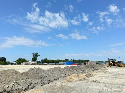 This media file depicts an outdoor construction or industrial site under a bright, blue sky with scattered white clouds. In the foreground, piles of dirt and gravel create a rough, uneven terrain, suggesting recent excavation or earthmoving activities.
The middle ground features a variety of objects and structures. To the left, several buildings with corrugated metal roofs and some windows are visible, surrounded by trees. Further to the right, a yellow backhoe loader, a piece of heavy construction equipment, is parked on the dirt. Behind the backhoe, there are stacks of materials including what appear to be large concrete blocks and spools of cable. Beyond these, arrays of solar panels are installed, suggesting a focus on renewable energy or a large-scale industrial operation. A blue shipping container and a group of red pipes are also present.
The scene is set during the daytime, likely late morning or early afternoon, given the bright sunlight and clear sky. The weather appears to be warm and sunny. The location cues, such as the style of buildings and the presence of solar panels, along with the provided context of Częstochowa, Poland, indicate an industrial or developing area. There is no discernible text visible in the image. The overall impression is one of an active or recently active work site.